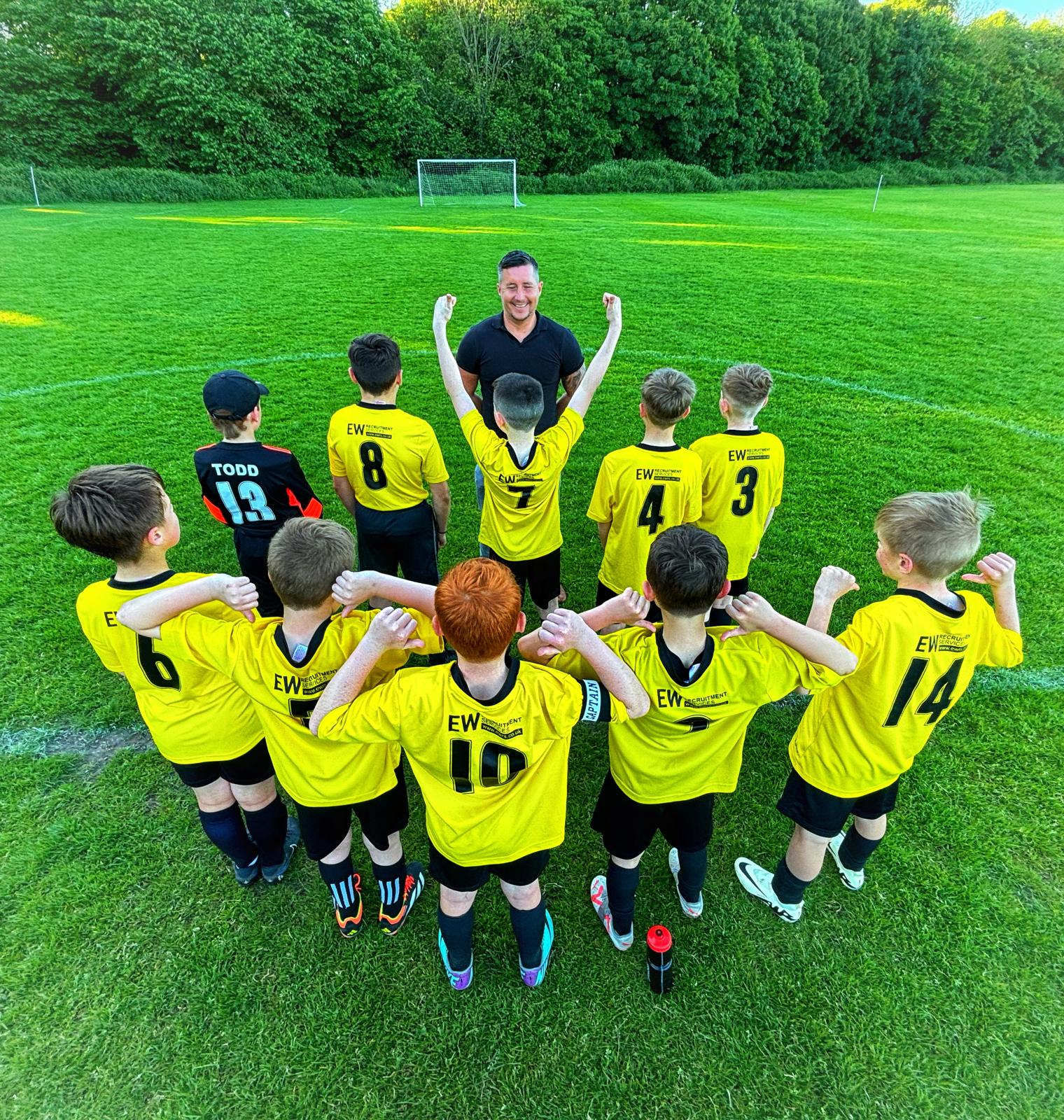 Grassroots football team with their backs turned to the camera, pointing to the EW Recruitment Services Logo on the back of their football shirts.
